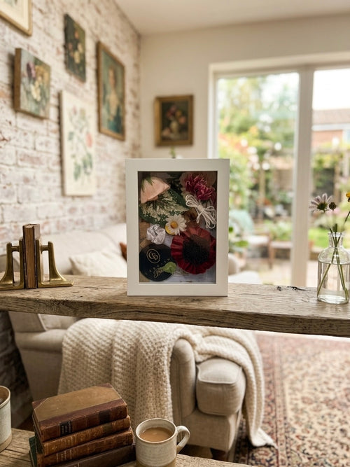 A white framed floral shadow box art piece displayed on a rustic wooden shelf in a cozy, sunlit living room with a brick accent wall, comfortable armchair, and garden view.