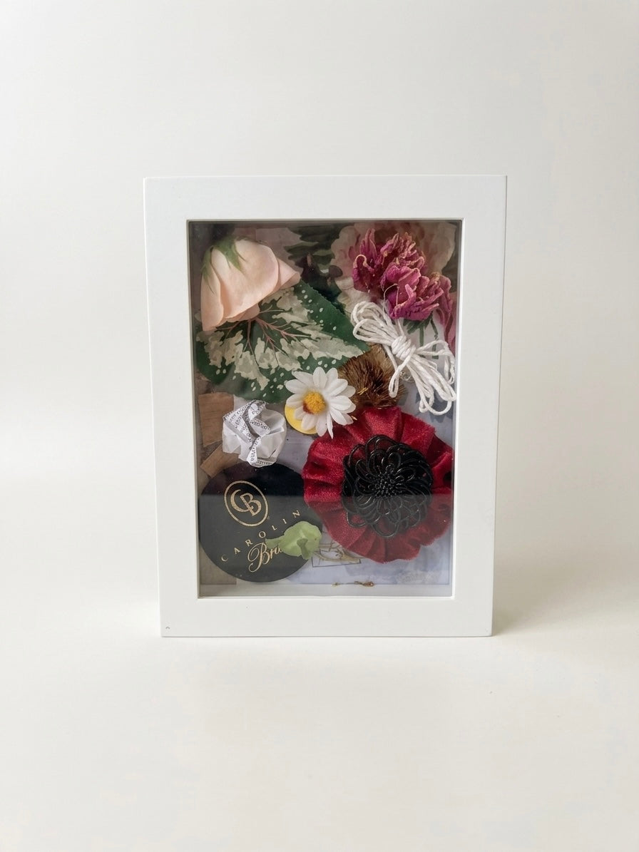 Handcrafted mixed media botanical shadow box artwork featuring a soft pink rose, a red textile flower, and green foliage layered inside a clean white rectangular frame against a bright, neutral white background.