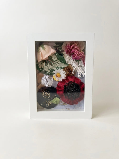 Handcrafted mixed media botanical shadow box artwork featuring a soft pink rose, a red textile flower, and green foliage layered inside a clean white rectangular frame against a bright, neutral white background.